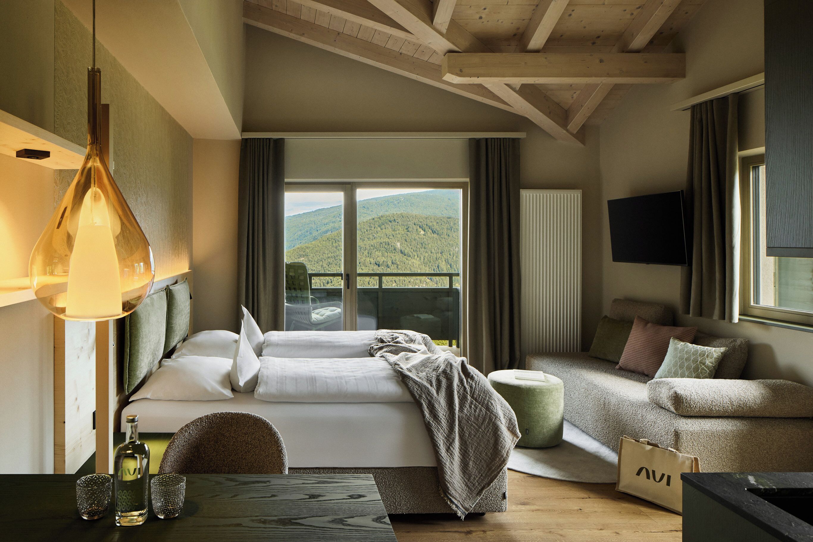 Un dormitorio con cama, sofá, televisor, una mesa con botella y vasos, y vista a una colina boscosa.