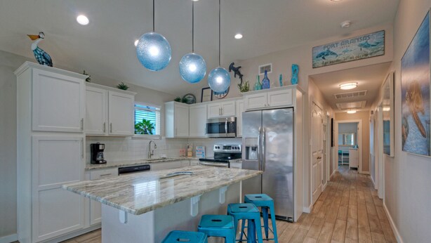A kitchen with white cabinets, a central island with stools, stainless steel appliances, and decorative items on the walls. - Private Pool + Lounge + Mins to Shore & Dining | Beach House | Tickle My Fansea (Port Aransas)