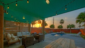 An outdoor patio with wooden furniture, string lights, and a view of palm trees and sunset. - Private Pool + Lounge + Mins to Shore & Dining | Beach House | Tickle My Fansea (Port Aransas)