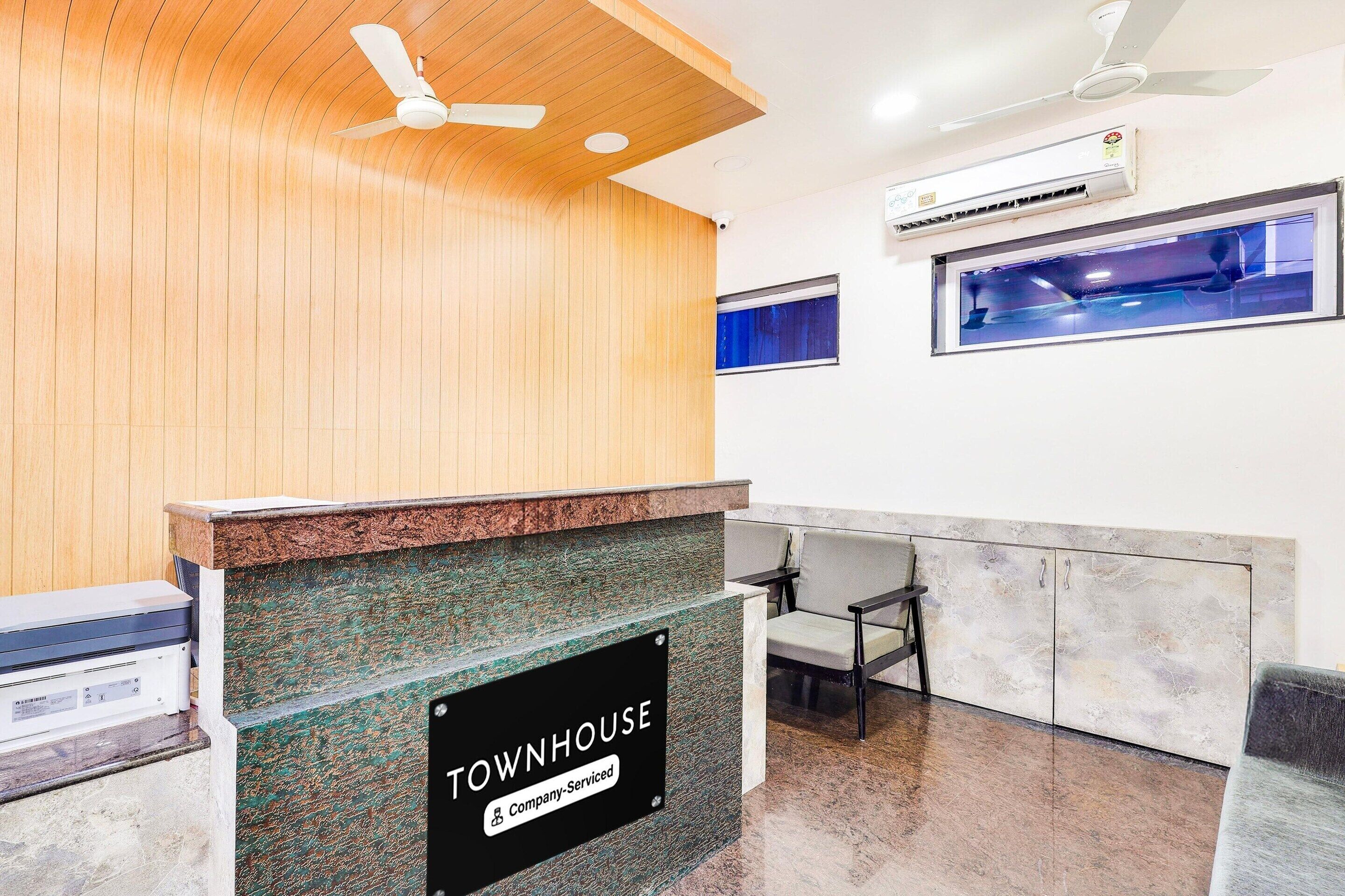 A reception desk with a sign reading 'TOWNHOUSE Company-Serviced,' a ceiling fan, an air conditioning unit, and a window with a view.
