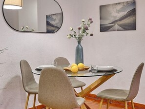 A dining area with a round glass table, grey upholstered chairs, a vase with flowers, and two framed pictures on the wall. - Apartamento Mes n C diz by Livework (Cádiz)