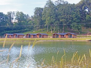 A row of wooden cabins with a lake in the foreground and trees in the background.