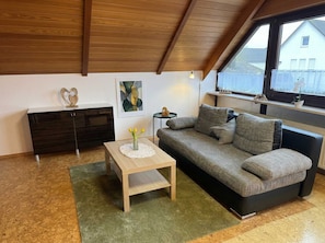 Un salon moderne avec un plafond haut, un meuble en bois foncé, un canapé gris, une petite table basse en bois et une fenêtre donnant sur une maison.