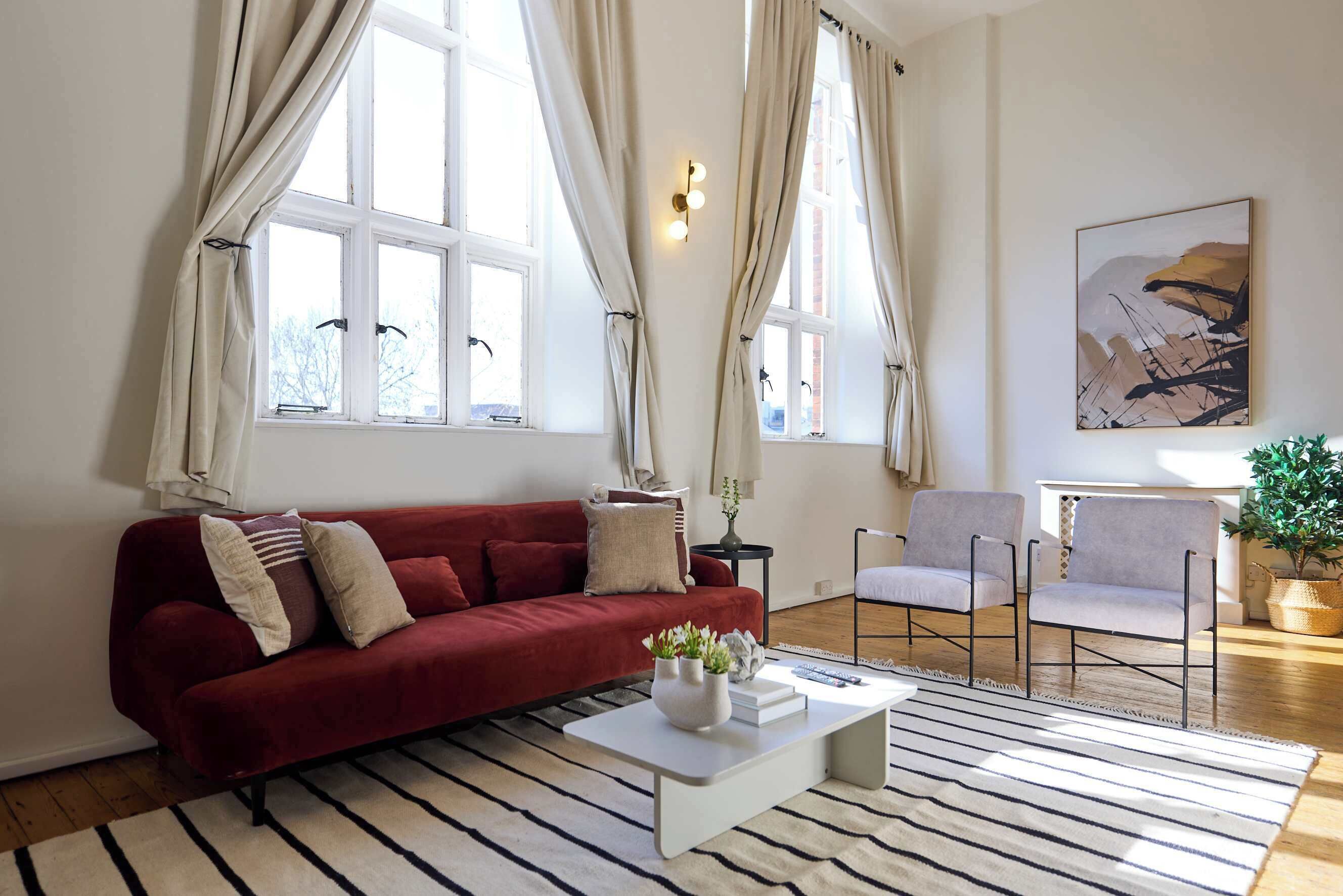 A living room with a red sofa, a white coffee table, and a striped rug. There are two windows with curtains, a painting on the wall, and a small table with a plant.