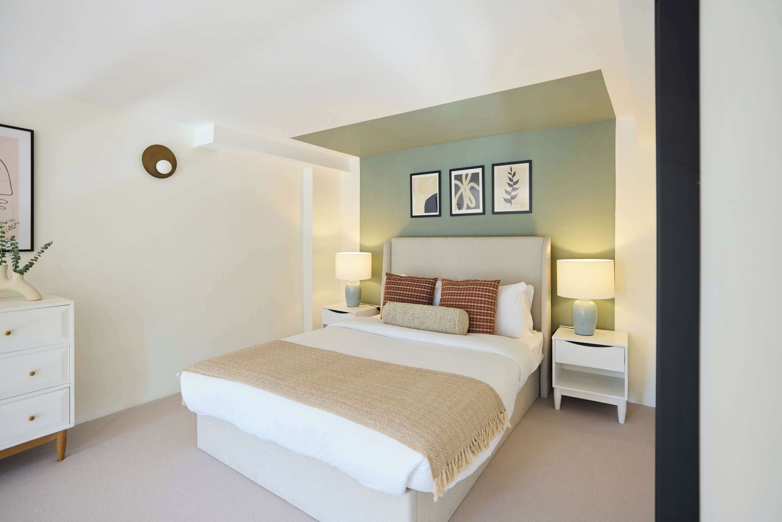 Une chambre à coucher comprenant un lit, des tables de chevet, une commode, une lampe fixée au mur et des œuvres d’art encadrées.