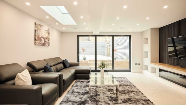 Een moderne woonkamer met een hoekbank, een glazen salontafel en een groot raam met zonwering.