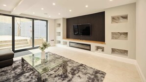 Een moderne woonkamer met een glazen salontafel, een flatscreen televisie bevestigd aan een houten paneel en een grote schuifpui naar een buitenruimte.