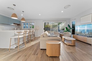 A modern living room with a kitchen area, wooden flooring, and a large window with a view. - Bay Breeze at Salamander Bay (Salamander Bay)