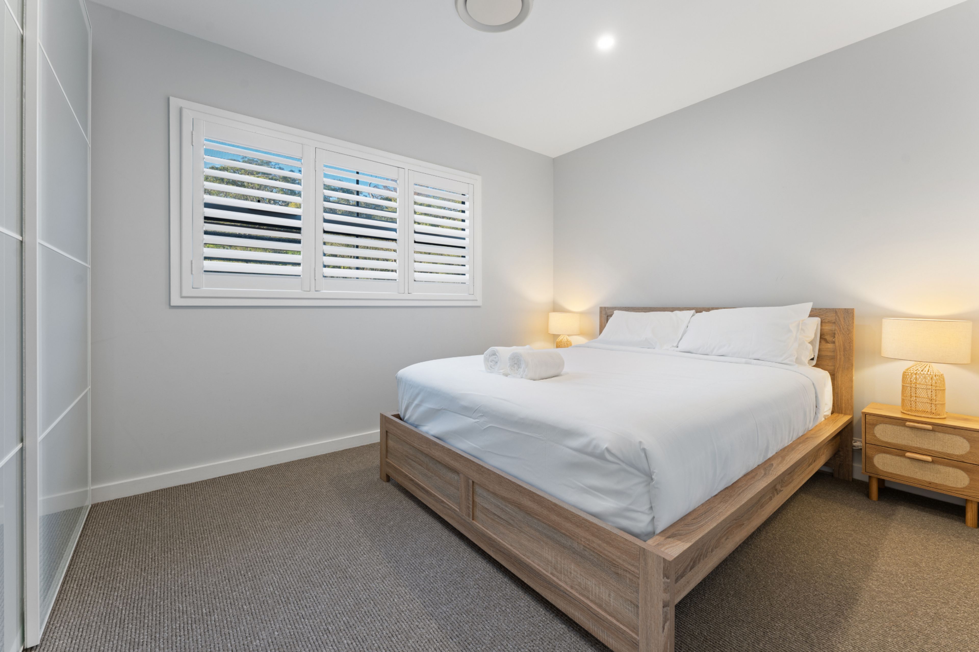 Een slaapkamer met een bed, twee nachtkastjes, een raam met zonwering en een vloerbedekking.