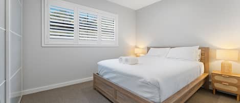 Une chambre à coucher avec un lit, deux tables de chevet, une fenêtre avec des stores et un sol recouvert de moquette.