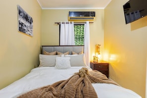 Une chambre à coucher comprenant un lit, une table de chevet avec une lampe, une fenêtre avec des rideaux, un climatiseur et un téléviseur fixé au mur.