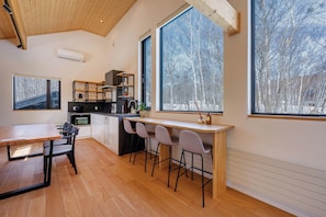 A modern kitchen with a wooden ceiling, a dining area with a table and chairs, and large windows with a view of trees.