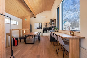 A modern interior with a wooden ceiling, a dining area with a bench, a kitchen with a stove, and a large window with a view of trees.