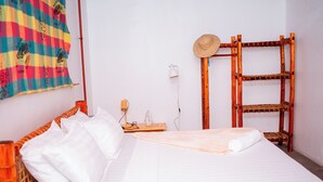 A bed with white linens, a wooden headboard, a straw hat, a woven shelf, and a colorful striped wall hanging.