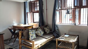 Quarto com sofá, mesa pequena, cadeira e janela com cortinas.