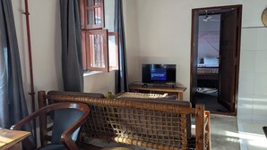 Quarto com televisão sobre suporte de madeira, janela com cortinas, porta para outro cômodo e cadeira de fibra.