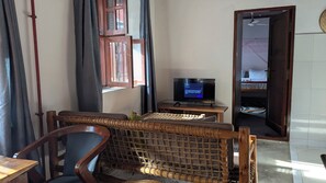 A room with a television on a wooden stand, a window with curtains, a door leading to another room, and a woven chair. - Kay's Place Train House Zanzibar Gallery (Zanzibar)