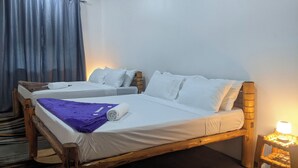 Uma cama de casal com estrutura de madeira, duas camas individuais, roupa de cama branca, um cobertor roxo e uma mesa de cabeceira com abajur.