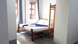 A single bed with a wooden frame, white bedding, and a blue blanket. There is a window with curtains, a patterned rug, and a wooden chair. - Kay's Place Train House Colonel tours (Zanzibar)