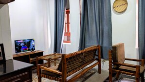 A room with a wooden sofa, a television, a woven chair, and a woven bed. - Kay's Place Train House Colonel tours (Zanzibar)