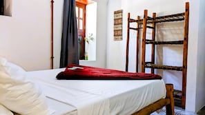 Ein Einzelbett mit weißer Bettdecke und roter Decke, ein hölzernes Etagenbett, ein Fenster mit Vorhang und eine Wand mit Wandteppich.
