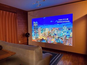 Una sala de estar moderna con un televisor de pantalla plana grande mostrando un paisaje urbano, piso de madera y un sofá gris.