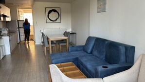 Ett modernt vardagsrum med en blå soffa, ett matbord med stolar och en köksdel med skåp och vitvaror.