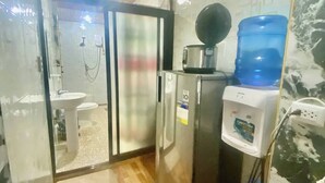 Una camera d'albergo con doccia, lavandino, frigorifero, dispenser d'acqua e una grande bottiglia d'acqua.