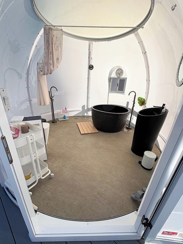 Une pièce circulaire et compacte, avec un intérieur blanc, une petite fenêtre, un lavabo blanc, une baignoire noire et un meuble de rangement blanc doté d’étagères.