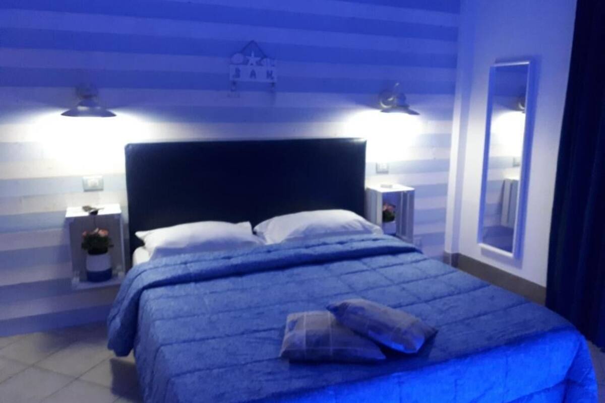 A bedroom with a bed, two bedside tables, a nightstand, a wall-mounted shelf, and a mirror.