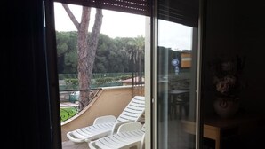 Una stanza con una porta a vetri scorrevoli che dà su un balcone con sedie in plastica bianca e vista su un paesaggio verde.