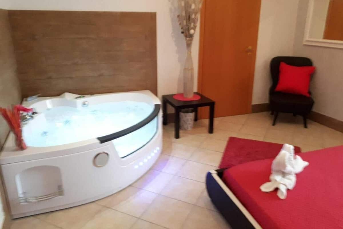 A room with a hot tub, a small table with a vase, a chair with a red pillow, and a bed with a red blanket.