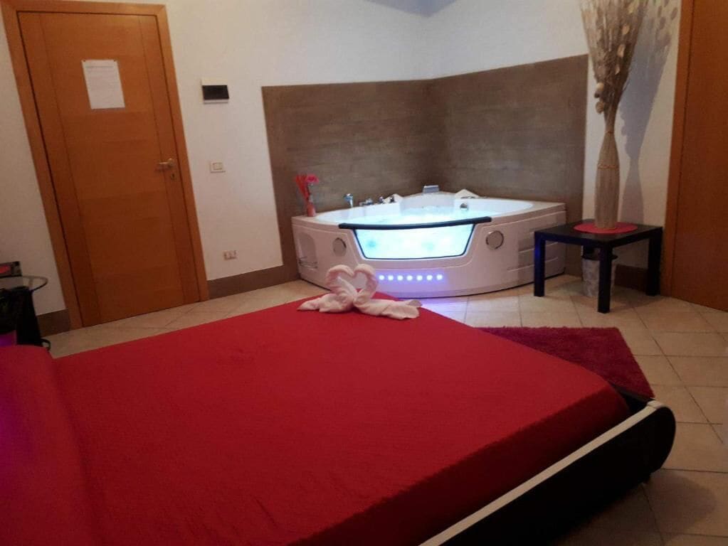 Un dormitorio con una colcha roja, una bañera blanca grande con luces LED, una mesita y una puerta.