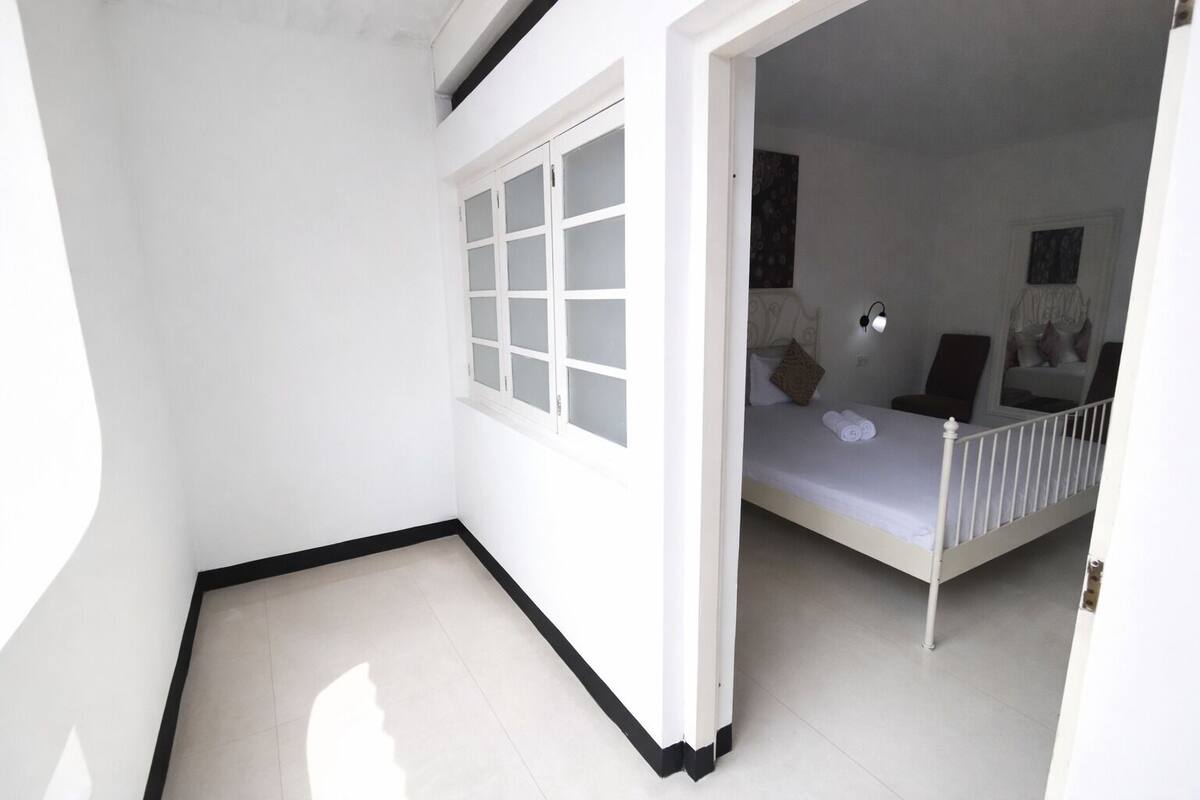 A bedroom with a white bed, a window with frosted glass, and a door leading to another room.