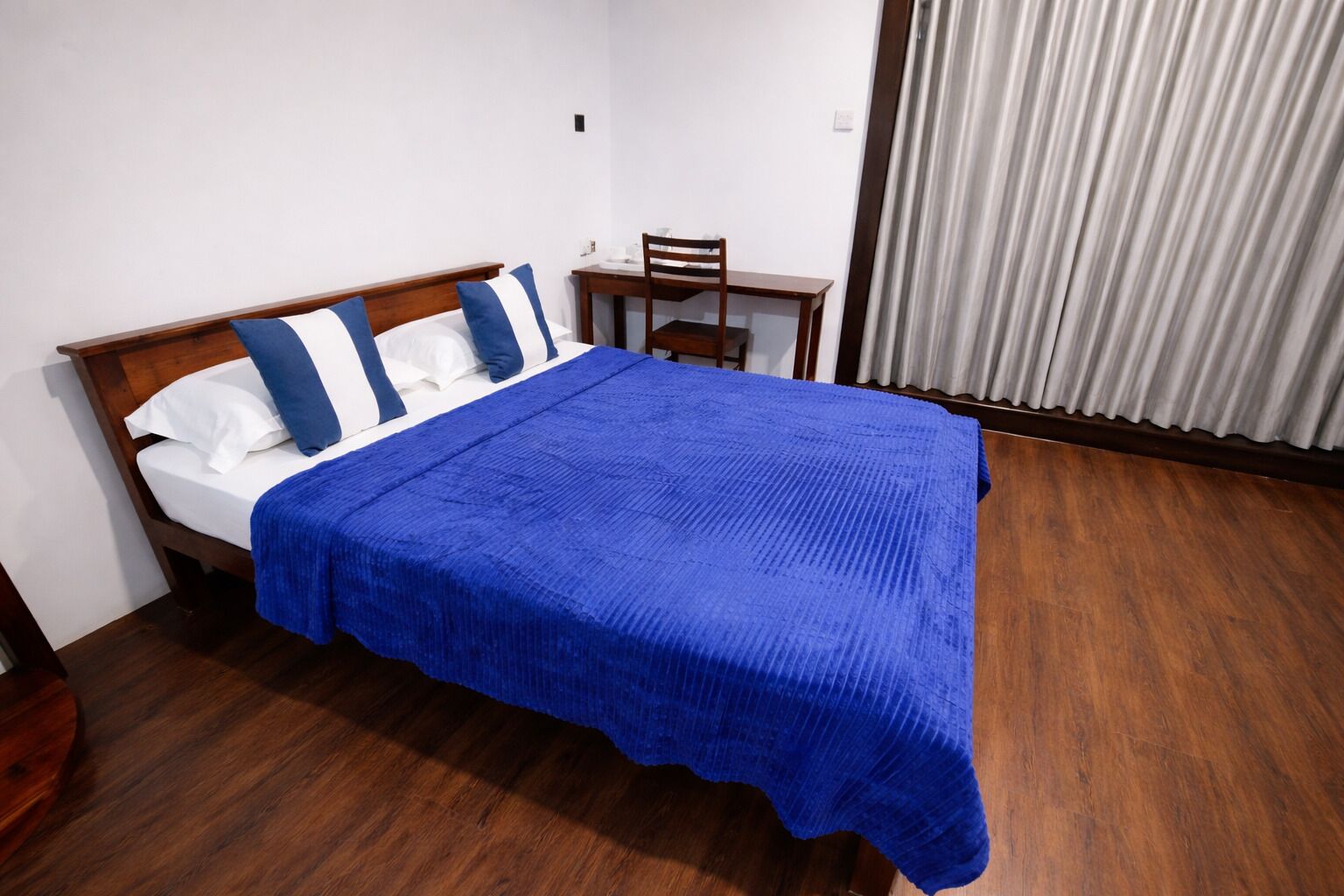 Una cama con manta azul, almohadas blancas y azules, cabecera de madera, una mesita de madera con una silla, y una ventana con cortinas.