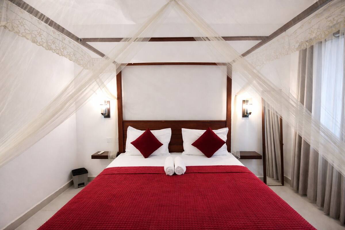 A hotel room with a large bed, white linens, red accents, a wooden headboard, and a canopy.