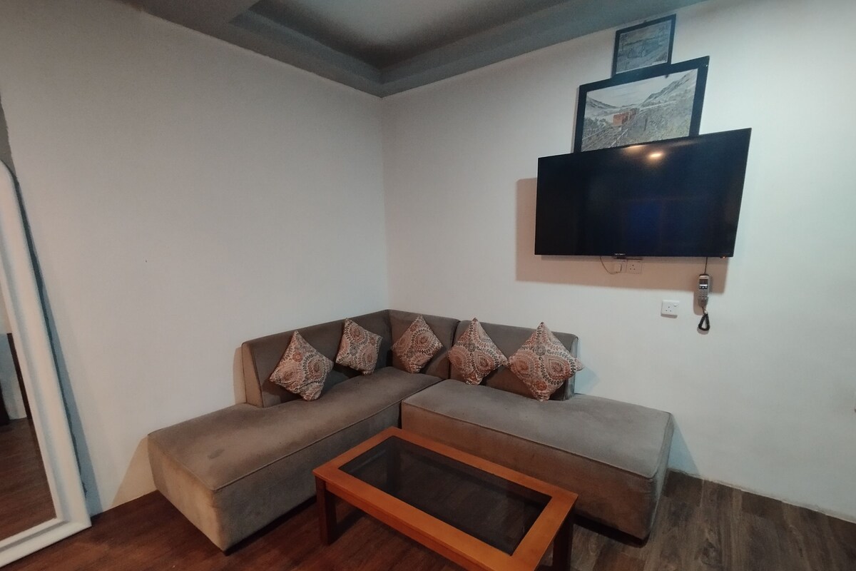A living room with a sectional sofa, a glass-top coffee table, a flat-screen TV mounted on the wall, and a telephone.