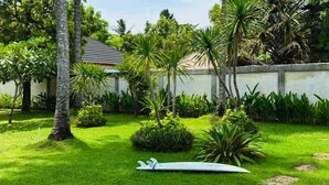 Un jardin bien entretenu, agrémenté d’une planche de surf blanche, de palmiers et d’un bâtiment doté d’un toit en tuiles.