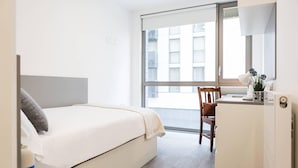A hotel room with a bed, a desk, a chair, a window, and a view of buildings outside. - Student Only Studio Chapter Lewisham (London)
