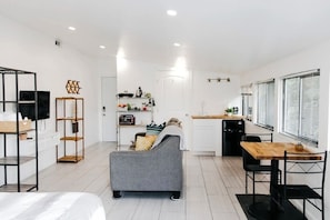 Una habitación moderna y luminosa con un sofá gris, una mesa de comedor de madera y una zona de cocina con refrigerador y fregadero.
