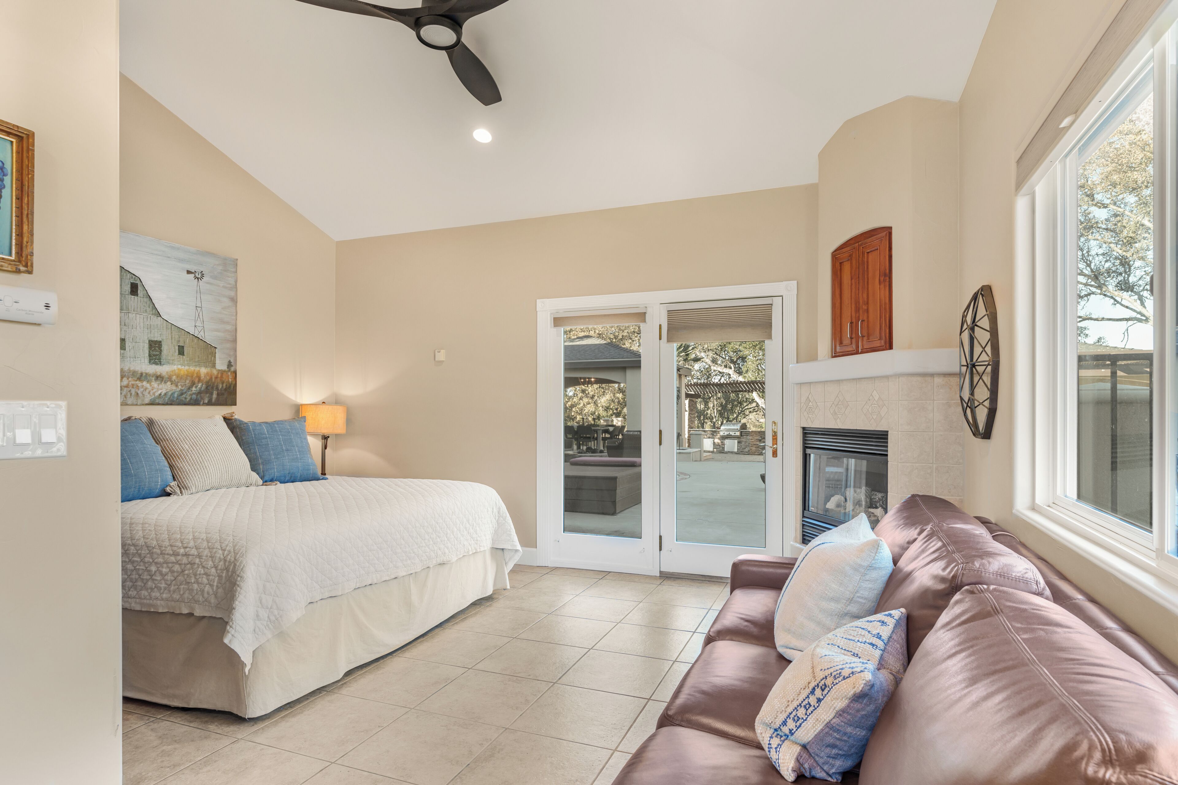 A bedroom with a bed, a brown leather couch, a fireplace, and a sliding glass door leading to an outdoor patio.