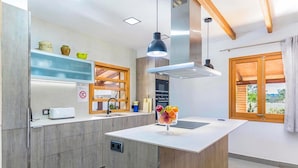 A modern kitchen with wooden cabinets, a central island, a built-in oven, and a window with a view. - Holiday Home "Yesero" with Mountain View, Pool, Wi-Fi & Garden (Illes Balears)