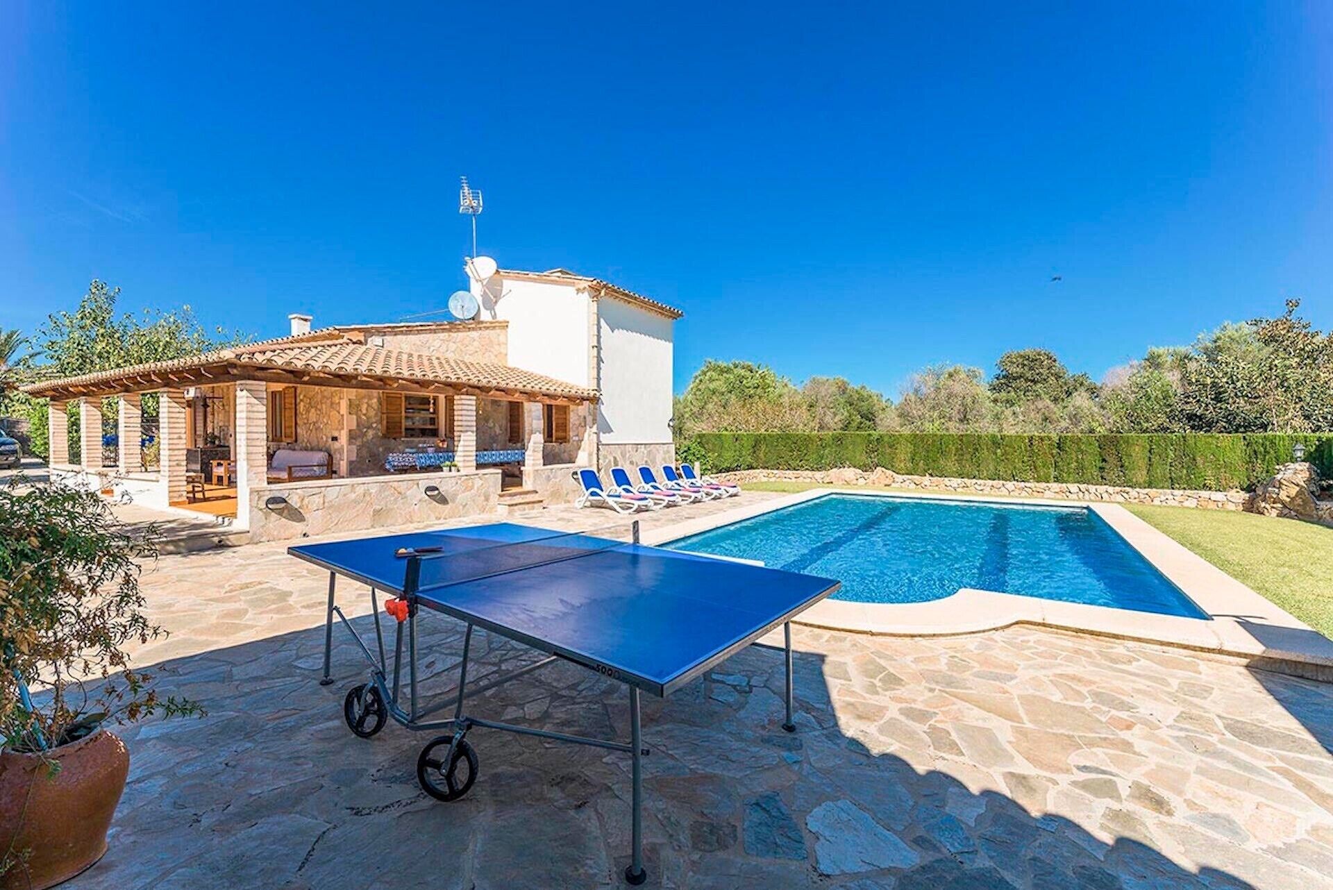 A pool area with a table tennis table, lounge chairs, and a building with a tiled roof.
