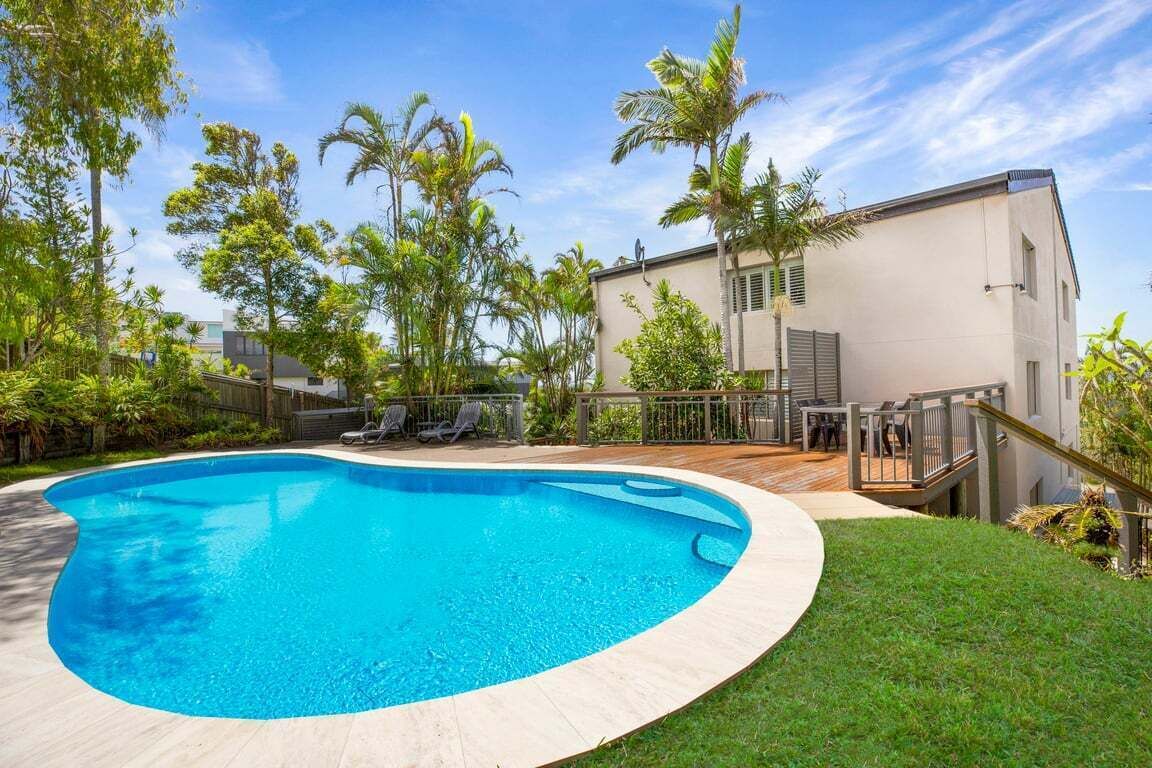A swimming pool with a wooden deck, surrounded by lush greenery and a modern building.