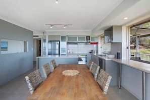 A modern kitchen with a central island, built-in appliances, and a dining area with a wooden table and woven chairs. - Captivating ocean views - Eagles Retreat  (Noosa)