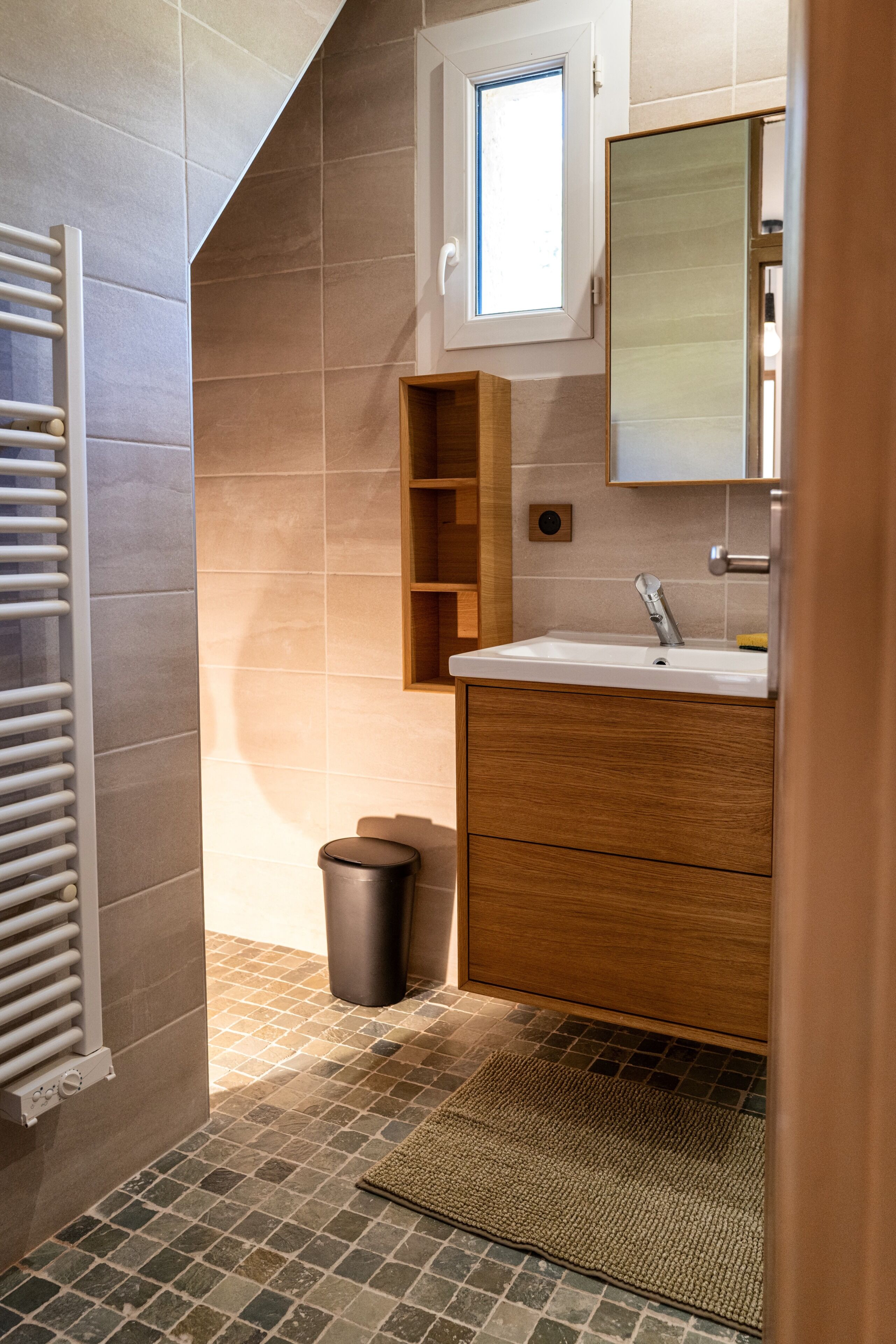 Un baño con suelo de baldosas, un lavabo, un espejo, un perchero para toallas y un cubo de basura.
