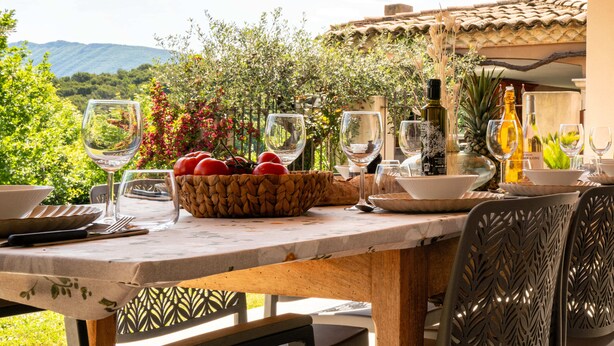 Mesa de refeições ao ar livre com copos de vinho, um cesto de tomates e uma garrafa de vinho, com um cenário de vegetação e montanhas.
