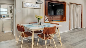 Comedor con mesa, sillas, un jarrón de flores, un letrero que dice 'WELCOME', un televisor y una cocina al fondo.
