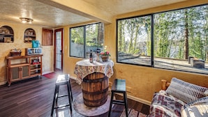 Ein rustikaler Innenraum mit einem Holzfässchen-Tisch, einer Couch und Blick auf den Wald durch große Fenster.