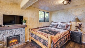 Ein Schlafzimmer mit Kamin aus Stein, einem Bett mit gemusterten Betttextilien, einem Nachttisch und einem an der Wand befestigten Fernseher.